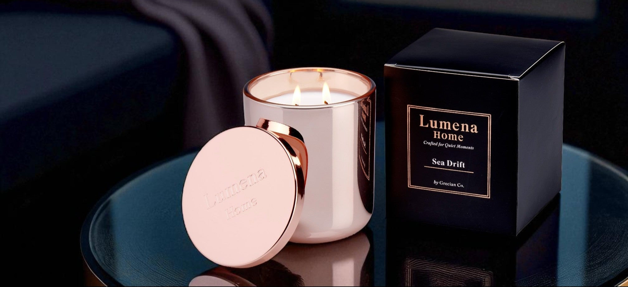 Rose gold candle and black box labeled 'Lumena Home' on a reflective surface with blurred lights in the background.